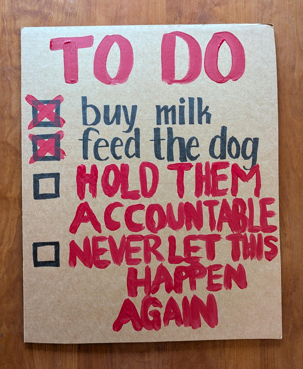 Handmade sign on a rectangle of cardboard, in black marker and red paint. “TO DO<br>☒ buy milk<br>☒ feed the dog<br>☐ hold them accountable<br>☐ never let this happen again”